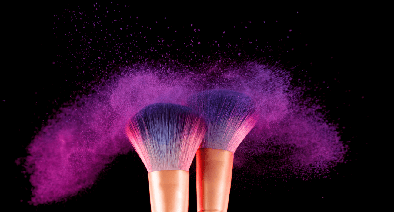 Two makeup brushes release purple powder into the air against a black background, creating a dynamic and artistic effect.
