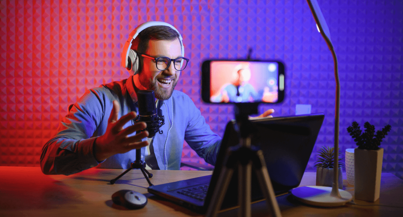 Creator con cuffie e microfono registra un video davanti a uno smartphone su treppiede, mentre parla sorridendo al laptop in uno studio illuminato con luci blu e rosse.